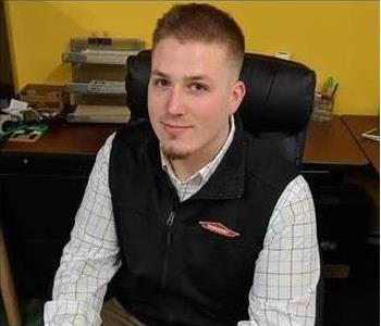 A man sitting in a chair at his desk wearing a sleeveless sweater bearing the SERVPRO logo